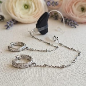 May include: A silver chain connects two small, round hoop earrings with clear, sparkling stones to a black hearing aid. The hearing aid is positioned near a soft, neutral background with flowers.