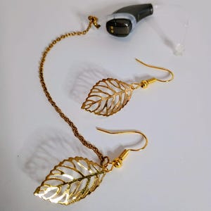 Puede incluir: Pendientes de hoja dorados con una cadena a juego, junto a un audífono negro y gris. Los pendientes presentan un delicado diseño de hojas caladas. El audífono tiene un tubo transparente.