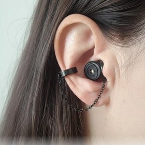 Pode incluir: Um ear cuff preto com uma corrente presa a um fone de ouvido preto. O ear cuff é uma faixa simples que envolve a orelha. O fone de ouvido é inserido na orelha. A corrente conecta o ear cuff e o fone de ouvido.