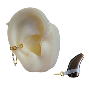 Puede incluir: Un pendiente tipo ear cuff dorado con una cadena y pequeñas piedras transparentes, conectado mediante un hilo fino a un audífono de color marrón oscuro. El pendiente está colocado en un modelo de oreja blanco, con el audífono al lado.