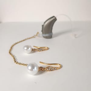 Puede incluir: Un par de pendientes dorados con grandes perlas blancas y pequeños cristales transparentes. Los pendientes están conectados por una delicada cadena dorada. También hay un audífono gris en la imagen.