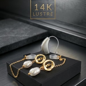 Puede incluir: Un par de dijes dorados para audífonos con perlas barrocas blancas y eslabones circulares irregulares, unidos por cadenas delicadas a bucles de silicona transparentes. El texto 14K LUSTRE es visible sobre la joyería en un fondo oscuro.