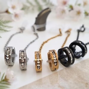 May include: A collection of six hoop earrings in silver, gold, and black tones, each featuring a row of clear embedded crystals. Each earring is attached to a thin metal chain, and the set includes a small dark grey hearing aid device.