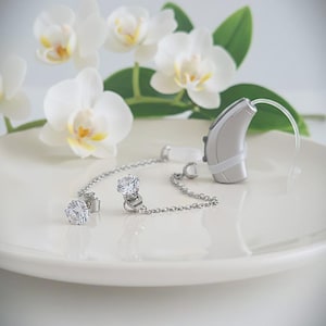 Può includere: Un accessorio per apparecchio acustico color argento caratterizzato da una sottile catenella metallica collegata a due ciondoli rotondi in zirconia cubica trasparente e sfaccettata. La catena si fissa al dispositivo con un anello in plastica.