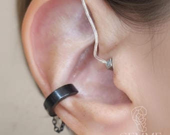 May include: A silver-toned ear cuff with a thin, curved wire extending upward and a small square accent piece, paired with a black ear cuff featuring a short, delicate hanging chain. The logo GEMME COMPANY is visible in the bottom right corner.