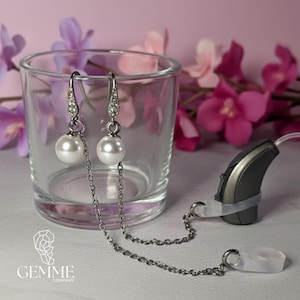 Peut inclure: Une paire de boucles d'oreilles en métal argenté avec des perles sphériques blanches et des cristaux transparents sur les crochets. Chaque boucle est reliée à une fine chaîne argentée fixée à une boucle en silicone pour appareil auditif.
