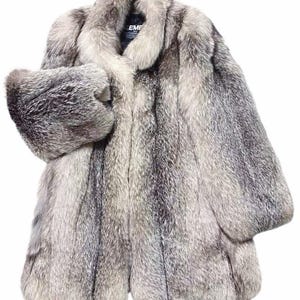 Puede incluir: Un abrigo de piel de longitud completa en tonos grises y plateados, con un manguito a juego. El abrigo tiene un cuello ancho y mangas largas. La etiqueta dice "EME".