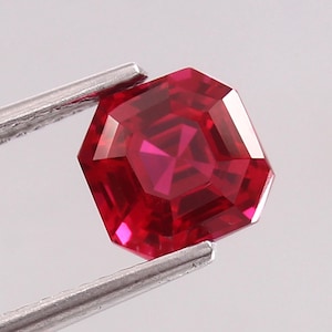 May include: A faceted, octagonal ruby gemstone held by silver tweezers. The gemstone is a deep red color with multiple facets that reflect light. The gem is a brilliant cut, showcasing its clarity and brilliance.