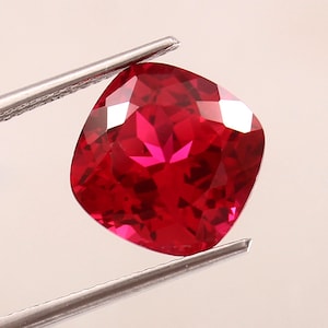 Puede incluir: Una gema roja de corte cojín sostenida por unas pinzas de metal plateado. La piedra presenta una superficie facetada que refleja la luz, creando reflejos carmesí profundos y escarlatas en su forma cuadrada y pulida.