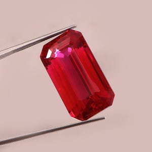 Op de afbeelding: Een gefacetteerde, rechthoekige robijn edelsteen, vastgehouden door een pincet. De edelsteen heeft een levendige rode kleur met meerdere facetten die licht reflecteren. De pincet is zilverkleurig en contrasteert met de kleur van de edelsteen.