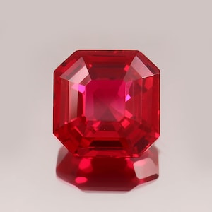 May include: A faceted, radiant-cut ruby gemstone. The gem is a deep, saturated red with a high degree of clarity, reflecting light and creating a brilliant sparkle. The gemstone is set against a neutral background, highlighting its color and cut.