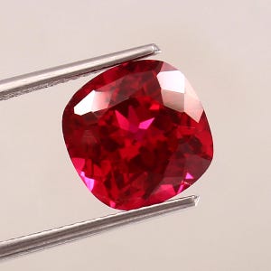Può includere: Un rubino taglio a cuscino, tenuto da pinzette d'argento. La gemma è di un rosso intenso con molteplici sfaccettature che riflettono la luce, creando un aspetto scintillante. La forma è quadrata con angoli arrotondati.
