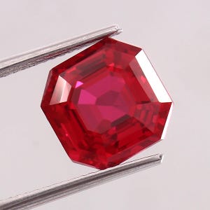 May include: A faceted, octagonal-cut ruby gemstone held by silver tweezers. The ruby is a deep, vibrant red color, with multiple facets reflecting light. The gemstone is a brilliant, eye-catching piece.