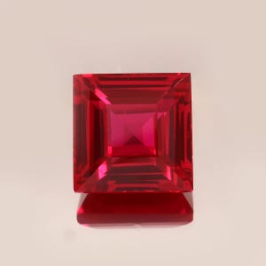 Puede incluir: Una piedra preciosa de rubí tallada en forma cuadrada y facetada. La gema es de color rojo vibrante con un alto grado de claridad, reflejando la luz de múltiples facetas internas. La piedra preciosa está sobre un fondo blanco.