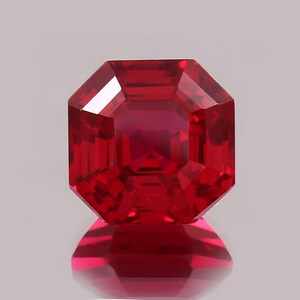 May include: A faceted, radiant-cut ruby gemstone. The gem is a deep, saturated red color with a high degree of clarity. The cut features a square shape with beveled corners, creating a brilliant display of light and color.