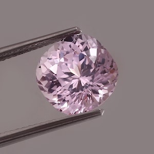 以下が含まれることがあります： 銀色のピンセットで保持された、クッションカットの淡いピンク色の宝石。宝石のファセットが光を反射しています。背景はニュートラルグレーです。宝飾品。