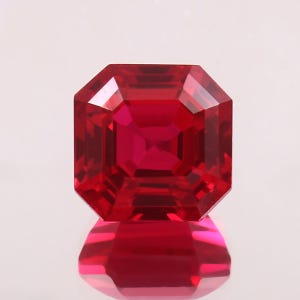 May include: A faceted, square-cut ruby gemstone. The gem is a deep, vibrant red color with a high degree of clarity. The facets reflect light, creating a brilliant sparkle. The ruby is set against a white background, with its reflection visible below.