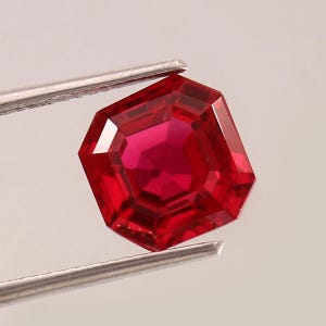 May include: A faceted, deep red gemstone held by silver tweezers. The gem has an octagonal shape with multiple facets, reflecting light. The color is a vibrant, saturated red, suggesting a ruby or similar precious stone.