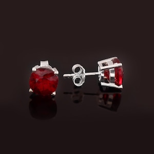 Könnte beinhalten: Ein Paar Ohrstecker mit leuchtend roten Edelsteinen, die in Silbermetall gefasst sind. Die rund geschliffenen Edelsteine werden von vier Krappen gehalten. Die Ohrringe werden vor einem dunklen Hintergrund gezeigt, der ihre Farbe und ihr Design hervorhebt.