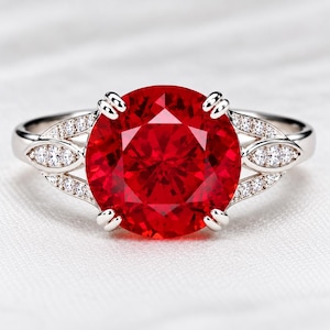 Può includere: Un anello color argento con una grande pietra centrale rotonda di colore rosso vivace, fissata da quattro griffe. La fascia è decorata su entrambi i lati da due sezioni a forma di foglia, ciascuna incastonata con tre piccole pietre bianche.