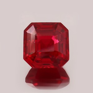 May include: A faceted, square-cut ruby gemstone. The gem is a deep, vibrant red color with multiple facets that reflect light. The gemstone is displayed on a reflective surface.