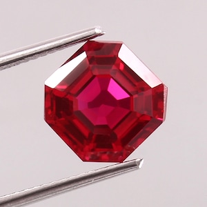 May include: A faceted, octagonal-cut ruby gemstone held by tweezers. The gem is a deep, vibrant red with a brilliant, reflective surface. The cut showcases the gem's internal structure and light play.