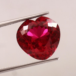 Pode incluir: Uma gema vermelha escura facetada em forma de coração é segurada por pinças prateadas. A gema tem múltiplas facetas que refletem a luz, criando reflexos brilhantes. A cor da gema é um vermelho rico e saturado, com um tom rosa claro.