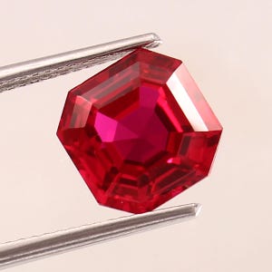Puede incluir: Una gema de rubí octogonal facetada, sujeta con pinzas plateadas. El rubí es de un color rojo intenso y saturado con múltiples facetas que reflejan la luz. La gema se encuentra sobre un fondo suave y neutro.