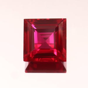 May include: A square-cut, faceted gemstone in a vibrant red hue. The gem has a symmetrical design with multiple facets that reflect light, creating a brilliant sparkle. The gemstone is set against a white background.
