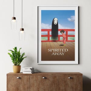 May include: Framed poster featuring a scene from the animated film "Spirited Away." The artwork depicts a young person in a red outfit facing a tall, dark figure with a white mask. The title "Spirited Away" is printed at the bottom.