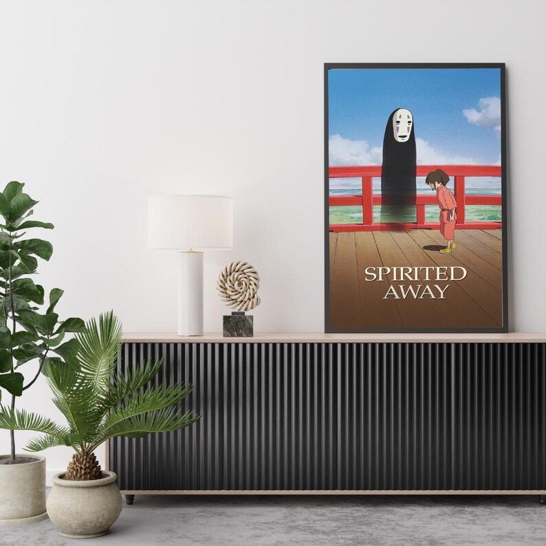 May include: Framed poster featuring artwork from the animated film "Spirited Away." The image shows a character in a red outfit standing on a wooden deck, with a tall, dark figure and the text "Spirited Away."
