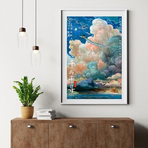 Op de afbeelding: Een ingelijste poster met een fantasie sc&egrave;ne met een draak in een bewolkte lucht, met Japanse tekst. Het kunstwerk gebruikt een levendig palet van blauw, oranje en groen. De poster wordt tentoongesteld op een houten kast.