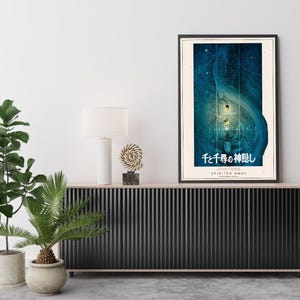 May include: Framed poster featuring a blue and teal illustration from the animated film "Spirited Away." The artwork includes Japanese text and the English title. The poster is displayed above a black cabinet with a white lamp and decorative objects.