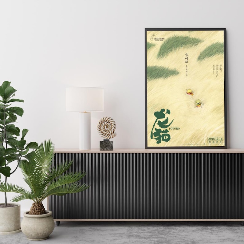 May include: A framed poster featuring a scene from the animated film "My Neighbor Totoro." The artwork showcases a field with green grass, two small figures, and Japanese text. The poster has a black frame and is displayed on a wooden cabinet.