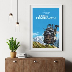 May include: A framed poster of Howl's Moving Castle, featuring the animated castle against a blue sky and mountain backdrop. The poster has the title in white text. The image is displayed on a wooden cabinet with a plant and hanging lights.