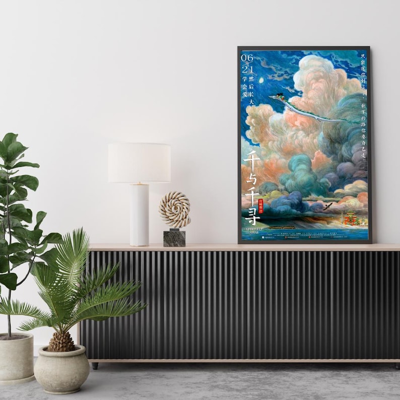 Op de afbeelding: Een ingelijste poster met een fantasierijke sc&egrave;ne van een draak in een bewolkte lucht, met Japanse tekst en data. Het kunstwerk wordt getoond op een zwarte kast, met een witte lamp en decoratieve objecten. De kleuren van de poster zijn levendig.