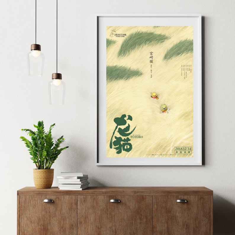 May include: A framed poster featuring the animated film "My Neighbor Totoro." The poster has a beige background with green grass-like shapes and two small figures wearing hats. The title "Totoro" is written in green Japanese characters.
