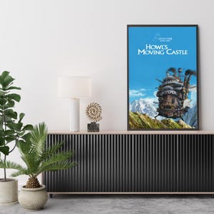 May include: Framed poster of Howl's Moving Castle against a blue sky with mountains. The artwork features the castle with smoke stacks, set on a green hillside. The title is in white, with Japanese text above.