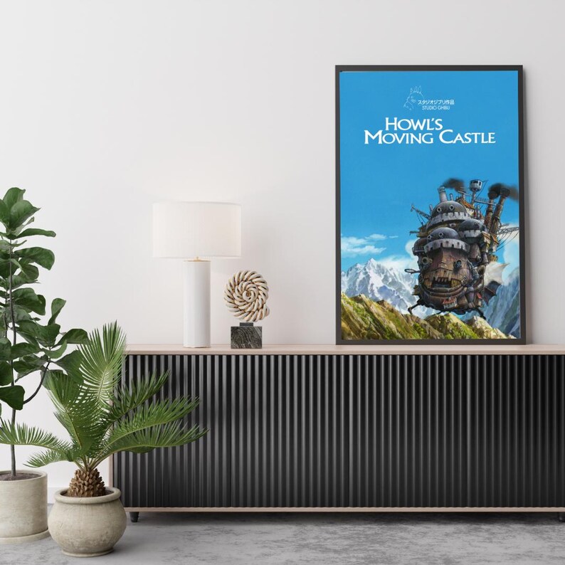 May include: Framed poster of Howl's Moving Castle against a blue sky with mountains. The artwork features the castle with smoke stacks, set on a green hillside. The title is in white, with Japanese text above.