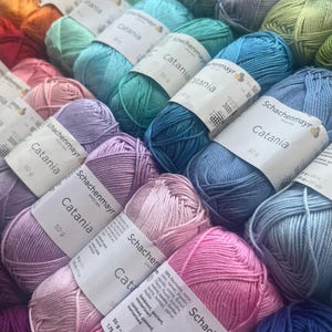 Peut inclure: Une collection de pelotes de fil de coton Schachenmayr Catania disposées en dégradé de couleurs incluant rose, violet, bleu, vert, jaune et orange. Chaque pelote de 50g porte une étiquette en papier blanc avec la marque et les détails.