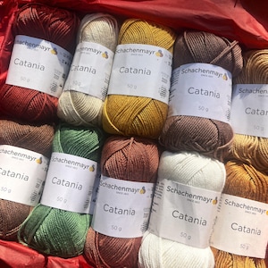 Peut inclure: Un ensemble de dix pelotes de fil Schachenmayr Catania de 50g dans des tons terreux variés, incluant rouille, crème, jaune moutarde, brun foncé, beige, vert olive et blanc, chacune avec une étiquette en papier blanc portant le nom de la marque.