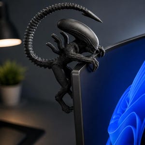 May include: A matte black figurine of a xenomorph creature with an elongated head and a curved, segmented tail. The figure is designed to perch on the top corner of a computer monitor, with its claws gripping the edge of the screen frame.