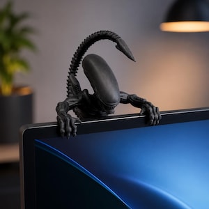 May include: A matte black Xenomorph figurine perched on the top edge of a computer monitor. The creature features an elongated head, sharp claws gripping the frame, and a segmented tail curving upward behind its back in a defensive posture.