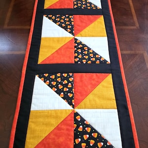 May include: A rectangular quilted table runner featuring a geometric pattern of triangles in white, orange, and mustard yellow, interspersed with a black fabric printed with small candy corn motifs, all framed by a solid black border and orange trim.