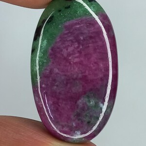 Puede incluir: Una piedra preciosa pulida de forma ovalada que presenta una mezcla vibrante de colores magenta intenso y verde bosque con inclusiones oscuras. La piedra tiene una superficie lisa y reflectante con un reflejo circular blanco central.