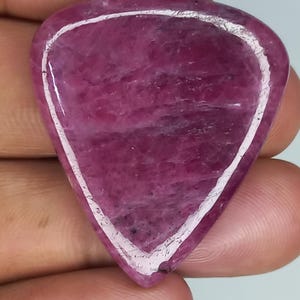 May include: A triangular-shaped, polished gemstone with a deep magenta and purple hue. The stone features a smooth, glossy surface with natural internal inclusions and a distinct, lighter-coloured border that follows the triangular perimeter.