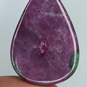 Può includere: Una gemma cabochon a forma di pera con un centro color magenta e viola intenso, caratterizzata da distinte inclusioni minerali verde scuro lungo i bordi. La superficie della pietra è lucida e liscia, con una consistenza naturale opaca.
