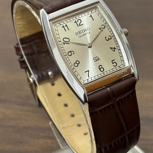 Puede incluir: Un reloj Seiko Quartz rectangular con caja plateada y correa de cuero marrón. La esfera del reloj es de color champán claro con números y agujas negros. La correa tiene un patrón de cocodrilo.