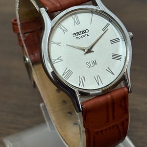 May include: A silver Seiko Quartz Slim watch with a white face and Roman numeral hour markers. The watch has a brown leather band with a crocodile pattern.