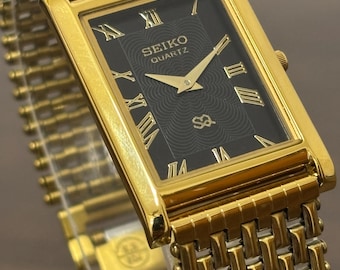 May include: A gold-toned Seiko Quartz watch with a rectangular black face and gold Roman numerals. The watch features gold hands and a gold link band. The watch face displays the Seiko Quartz logo. The watch is a classic design.